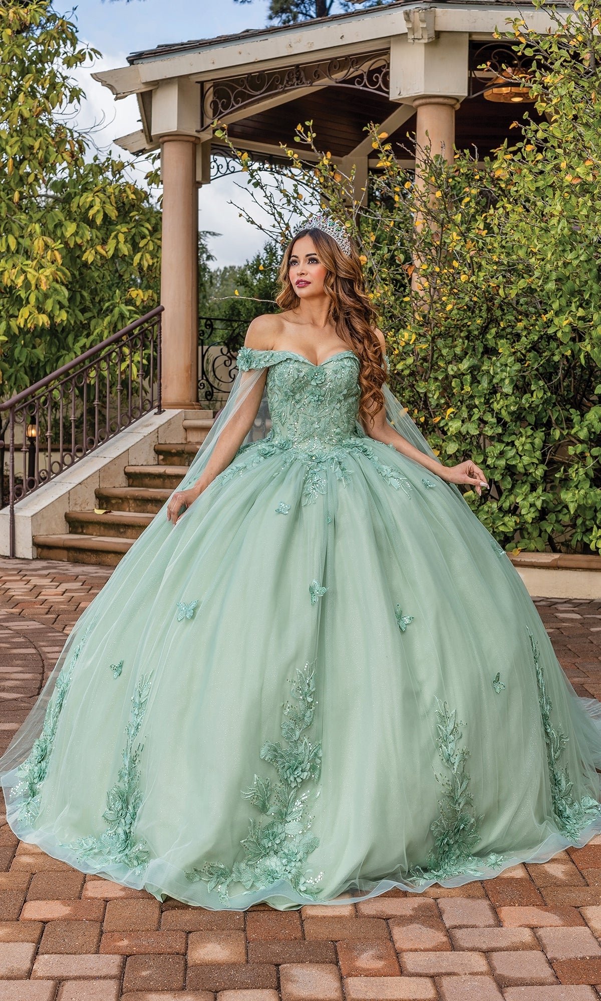 A woman stands in front of a gazebo on a brick pathway, wearing the mint green Quinceanera Dress 1853 By Dancing Queen with off-the-shoulder floral appliqué.